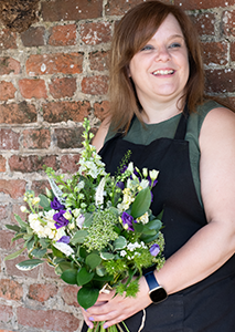 smiling light brown haired lady wearing a black apron and sleeveless green top holding a bouquet of white and purple flowers
